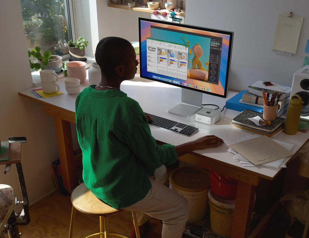 Person working on a Mac Mini M4 at a desk with plants and art supplies, using a large monitor for design tasks.