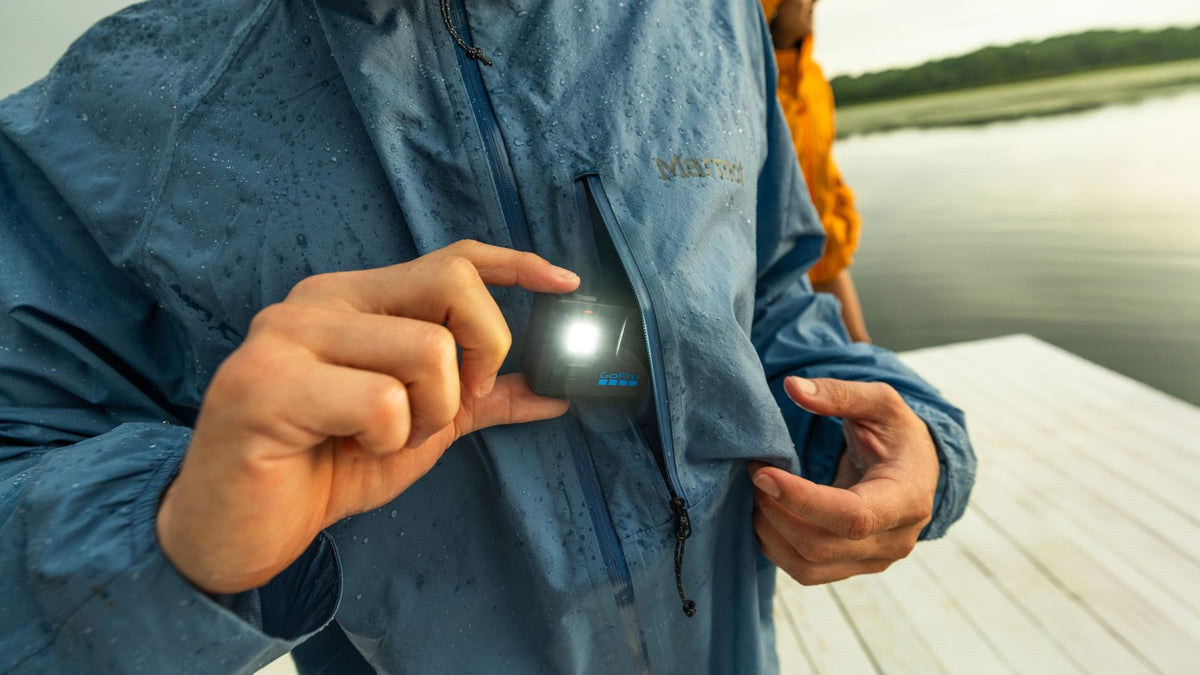 alAsil GoPro LIT HERO action camera with built-in light held by a person in a rain jacket near a lake