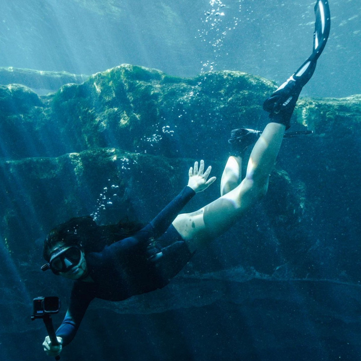 alAsil diver using a GoPro MISSION 1 cinema camera to film underwater in a rocky, deep blue aquatic environment.